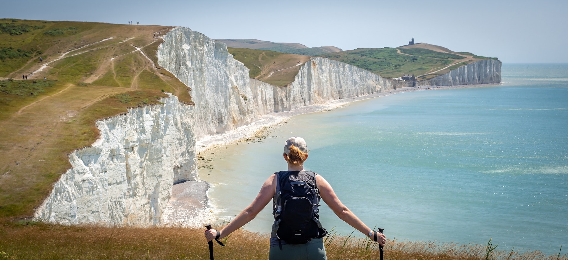 A hiker stands on a trail looking ahead at white chalk cliffs that rise and fall along the coastline on the South Downs Way